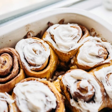 Golden-brown pumpkin cinnamon rolls arranged in a baking dish, topped with rich cream cheese frosting and a sprinkle of cinnamon.  