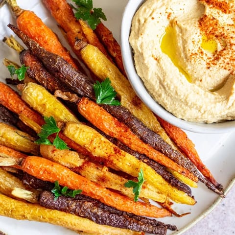 A close-up of golden roasted rainbow carrots and hummus, showing drizzled olive oil and fresh parsley on a serving platter.