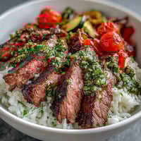 Sizzling grilled steak slices lie over fluffy white rice and colorful roasted vegetables, drizzled with fresh chimichurri sauce in a vibrant bowl.  